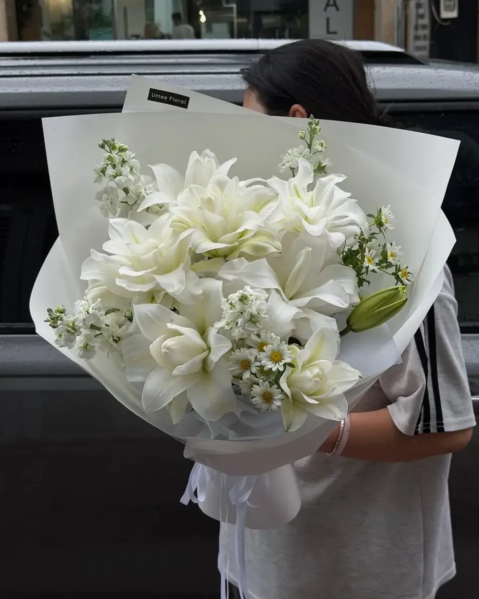 Bó hoa trắng gồm ly, hồng và cúc nhỏ gói giấy trắng, một người cầm trước nền tối