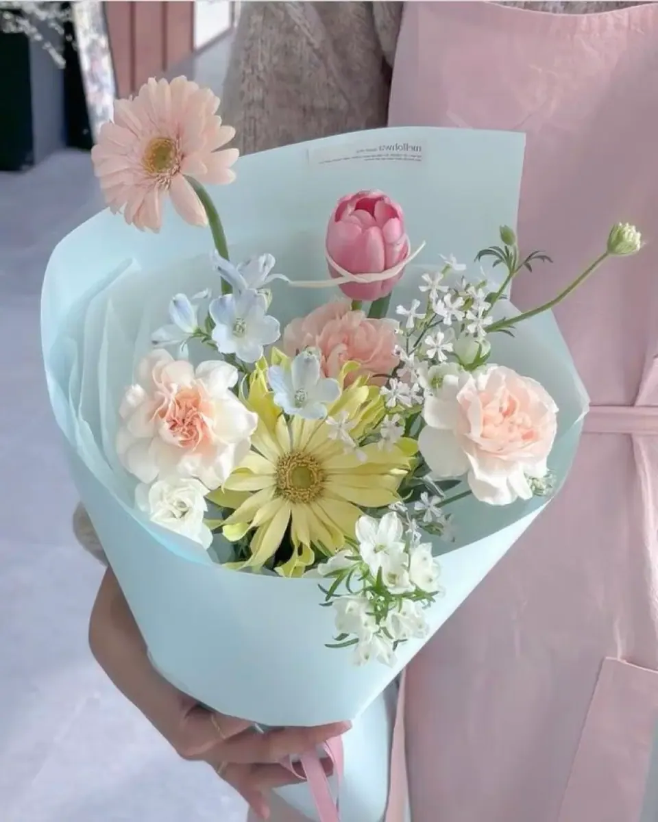 Bó hoa mềm nhiều màu hồng, vàng, trắng gói giấy xanh bạc hà thắt nơ, người cầm trước mặt