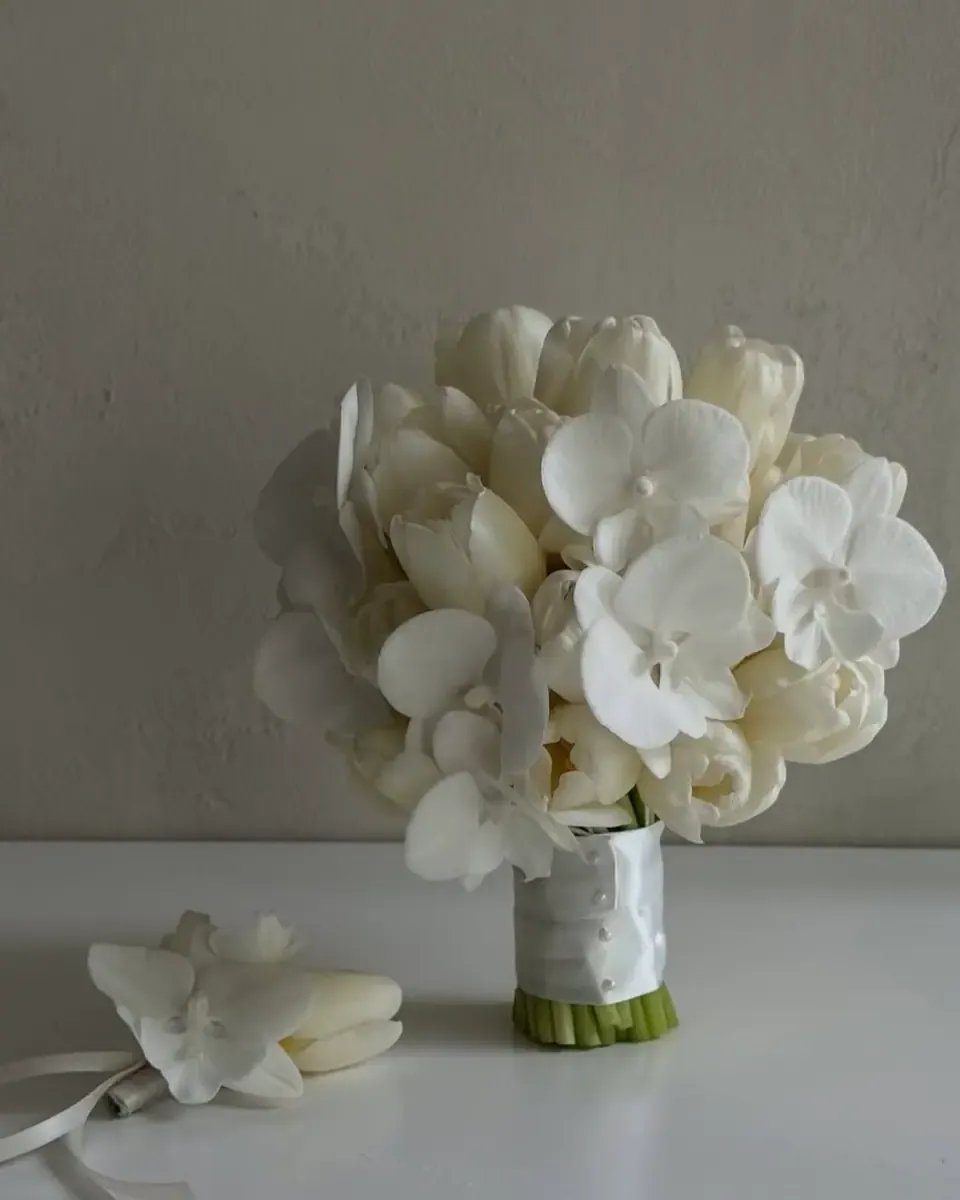 Bó hoa lan trắng và tulip trắng trong bình thấp, bên cạnh là một bông lan cài áo