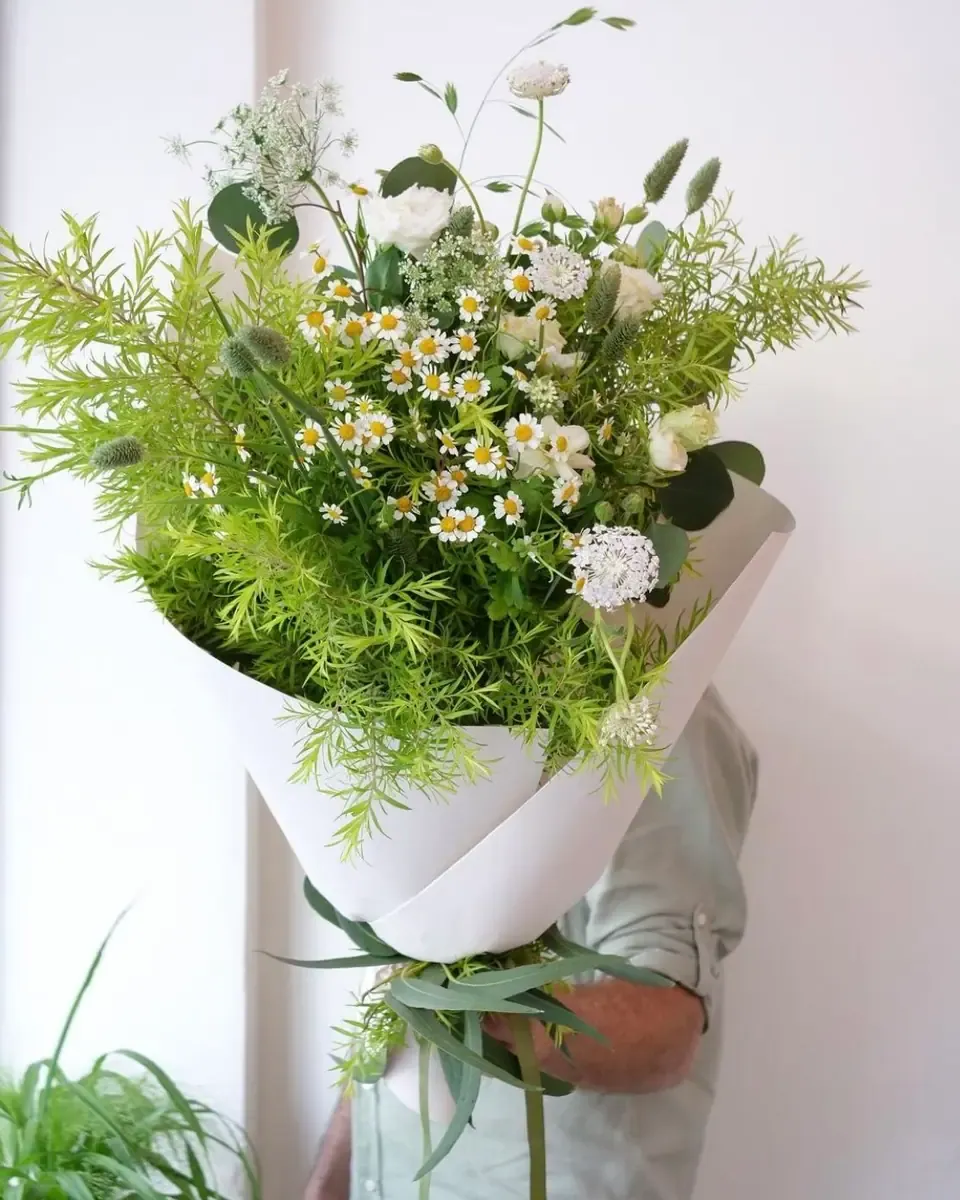 Bó hoa gói giấy trắng với lá xanh mảnh, cúc nhỏ trắng vàng và hoa trắng, một người cầm phía sau