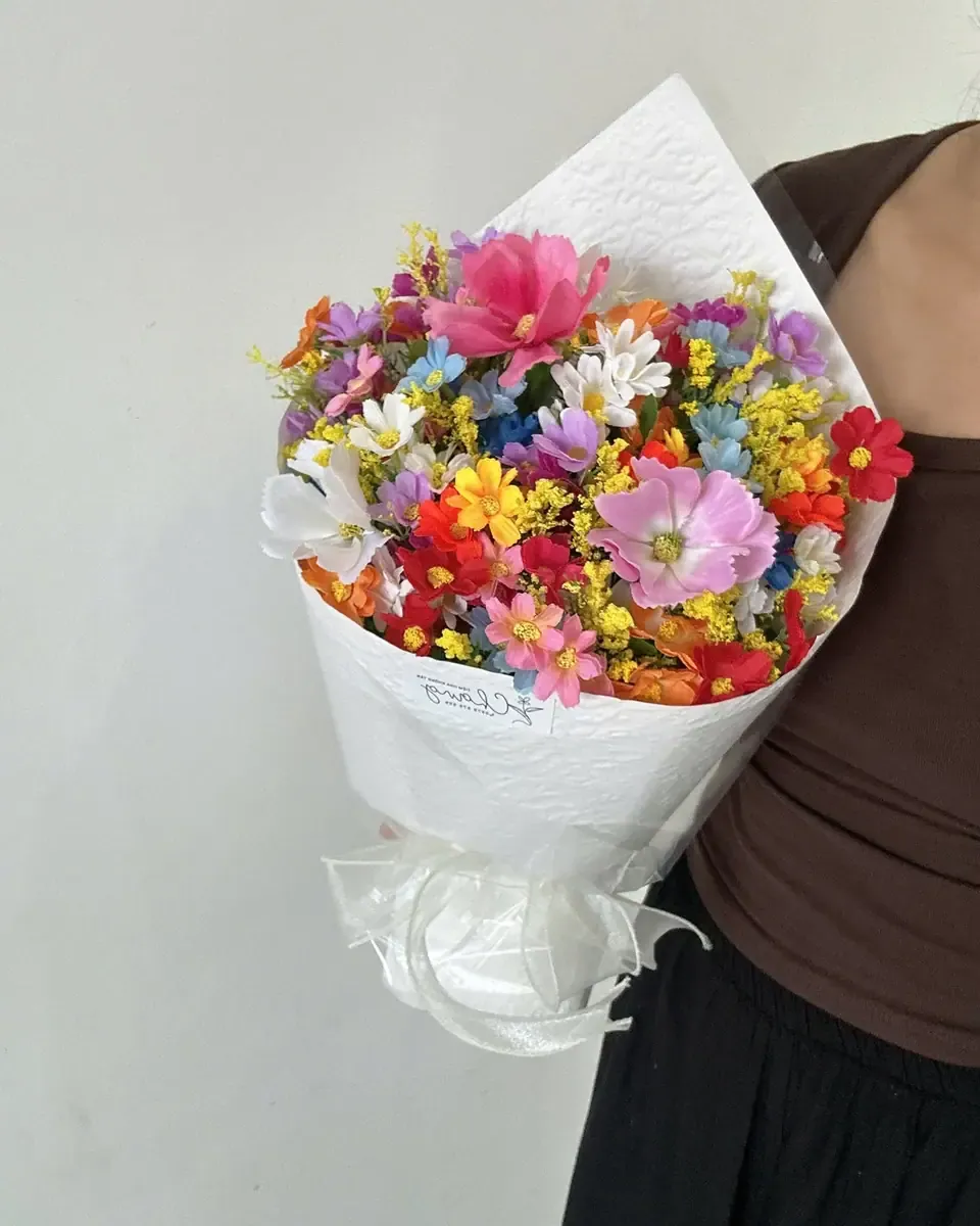 Bó hoa nhỏ nhiều sắc đỏ, vàng, hồng gói giấy trắng, cầm trước bức tường sáng