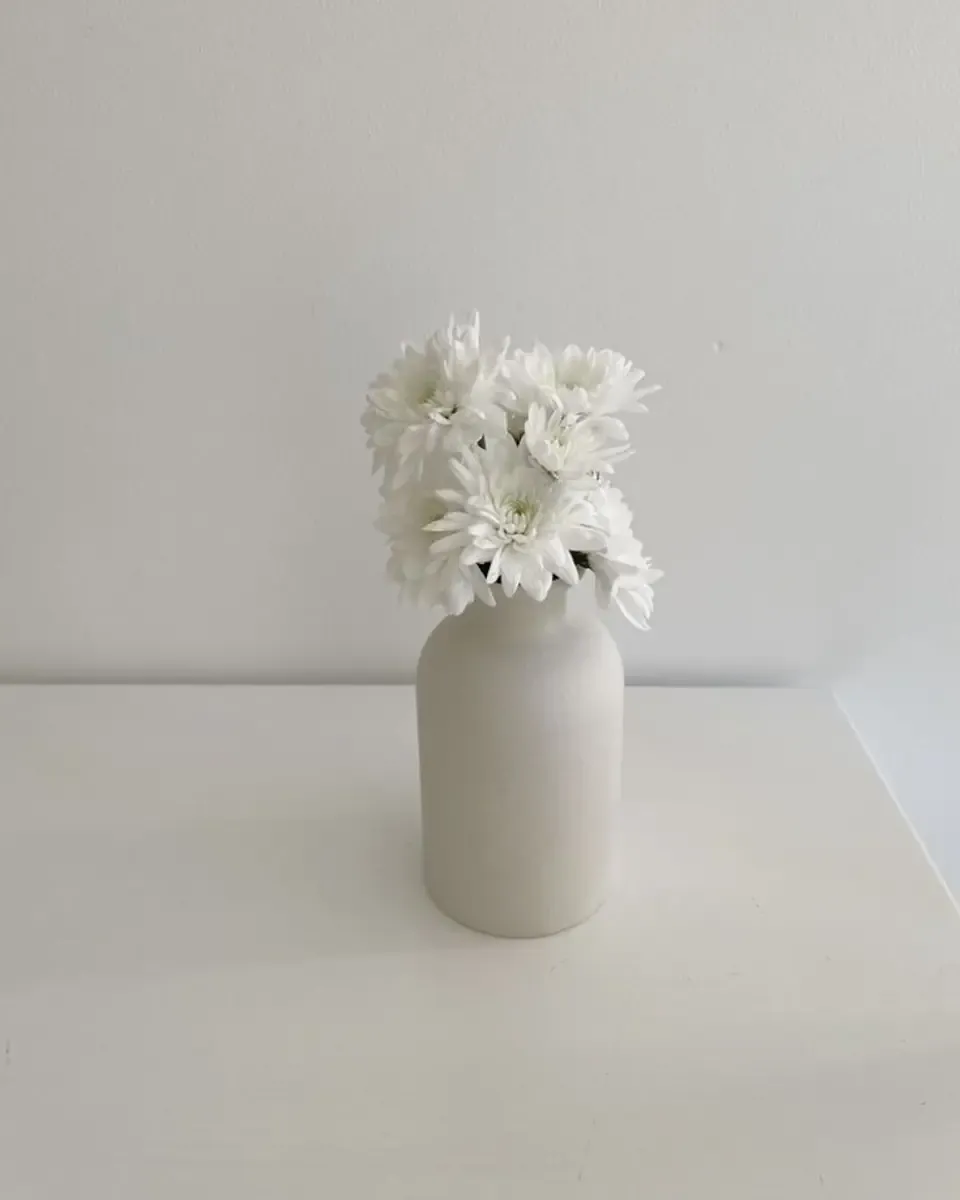 Bình sứ trắng dáng chai cắm cụm hoa cúc trắng đặt trên mặt bàn trắng gọn gàng