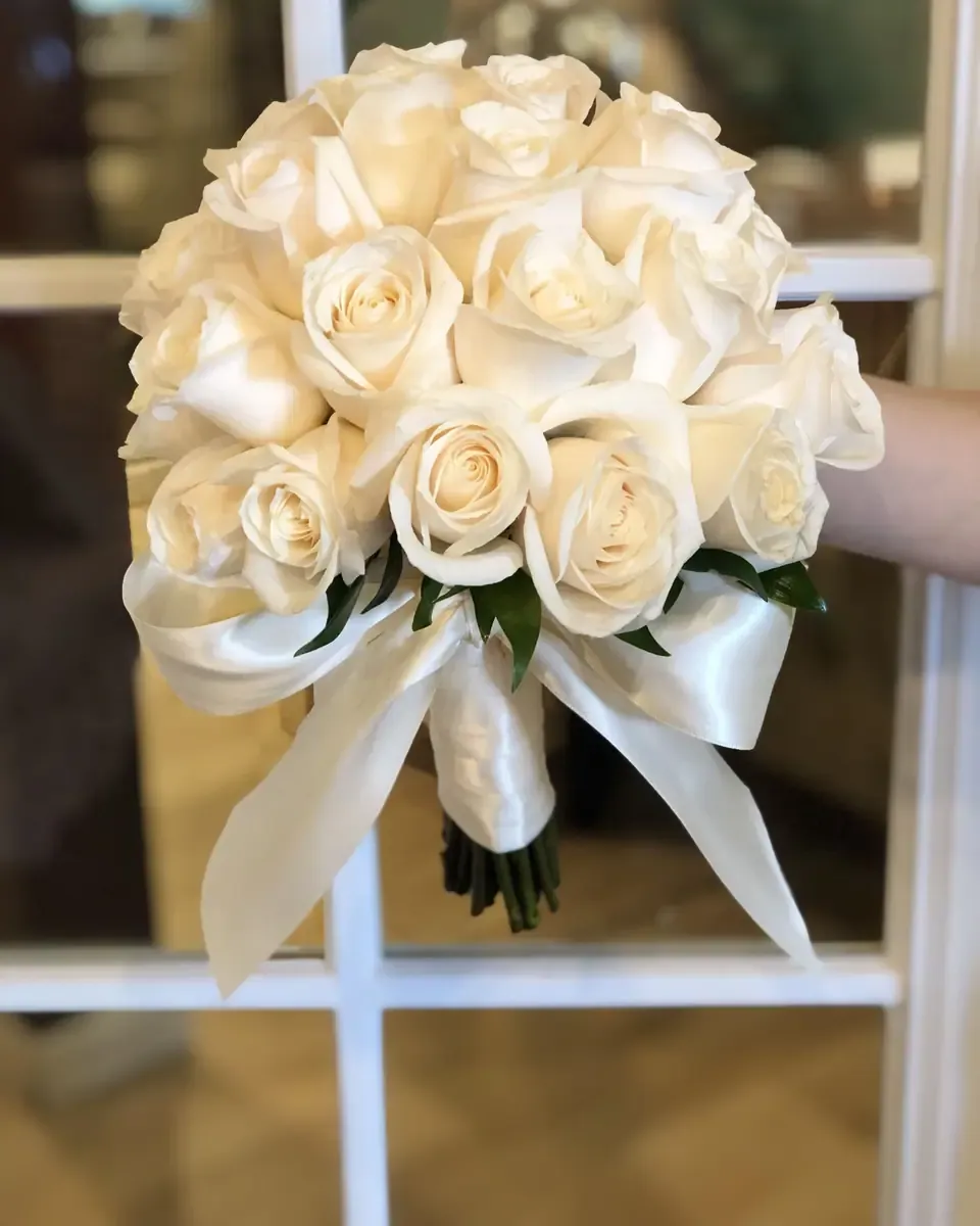 Bó hoa hồng trắng tròn dày buộc nơ lụa trắng lớn trước khung cửa kính