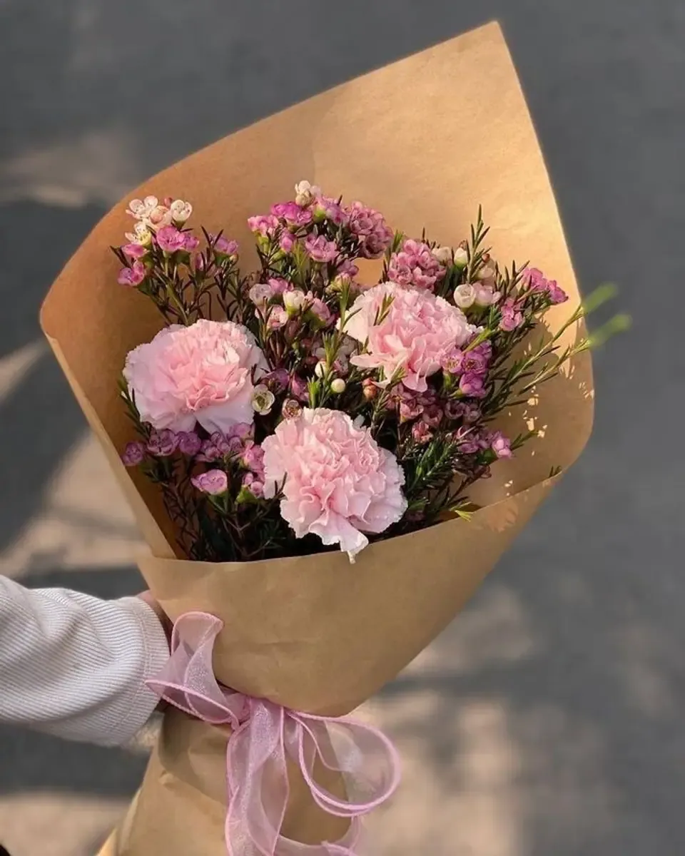 Bó cẩm chướng hồng cùng hoa sáp tím gói giấy kraft nâu, được cầm trên tay