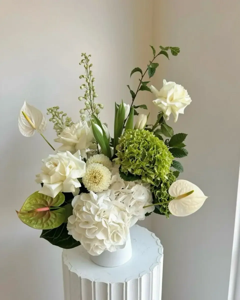 Bình gốm trắng nhỏ trên bục gân cắm hoa hồng trắng, cẩm tú cầu xanh, hoa môn trắng và lá xanh