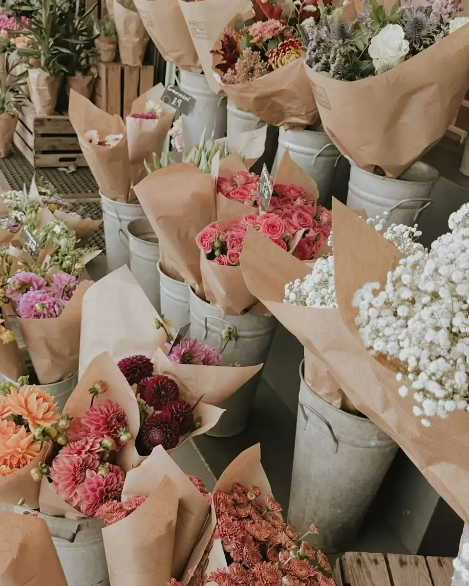 Nhiều bó hoa hồng, cẩm chướng và hoa trắng gói giấy kraft cắm trong xô tại gian hoa