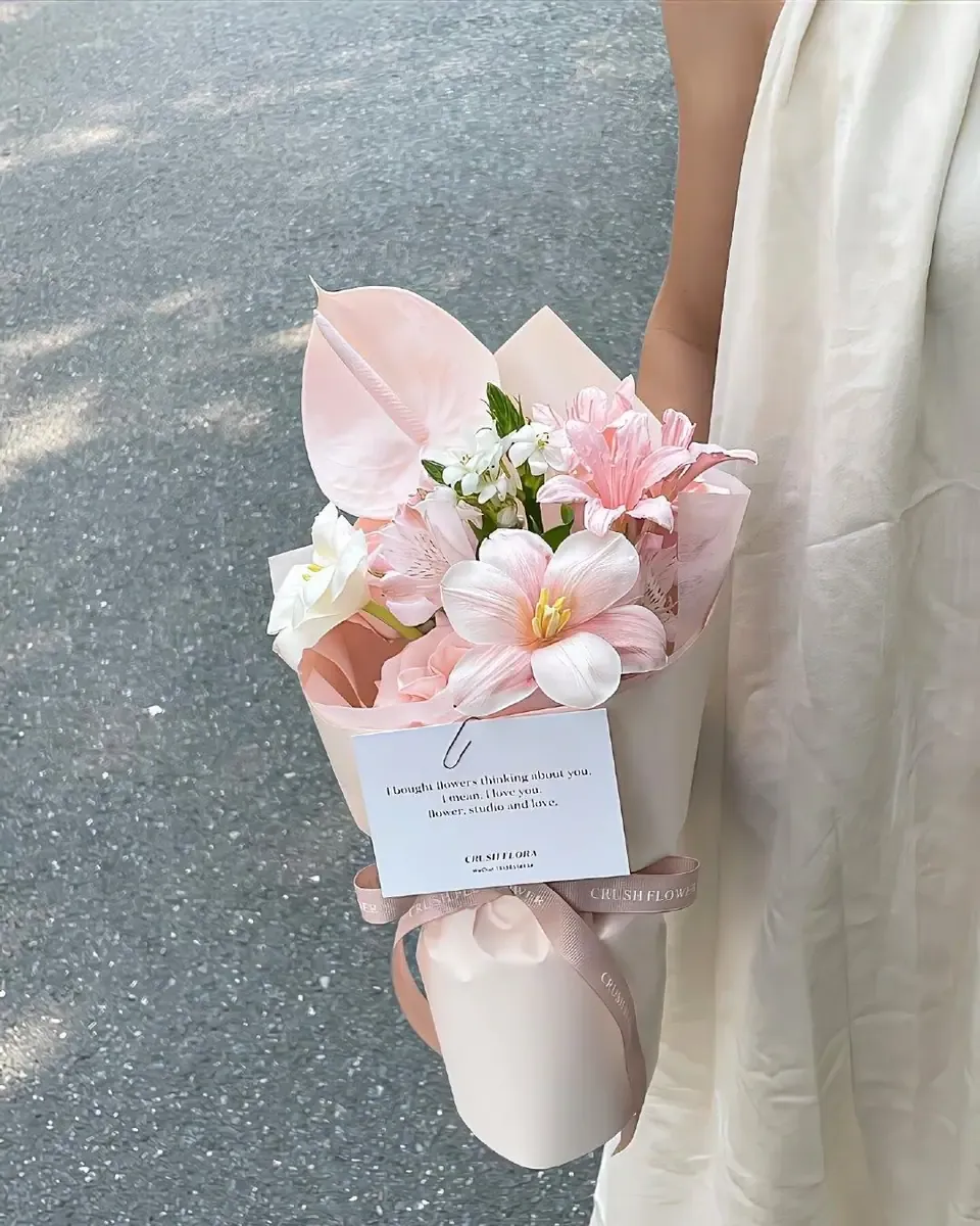 Bó nhỏ hoa môn hồng, ly hồng nhạt và hoa trắng gói giấy hồng kem, một tay cầm