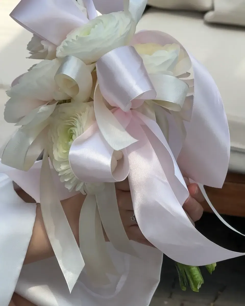 Bó hoa trắng nhiều ruy băng satin lớn được một bàn tay cầm, nền sáng nhẹ