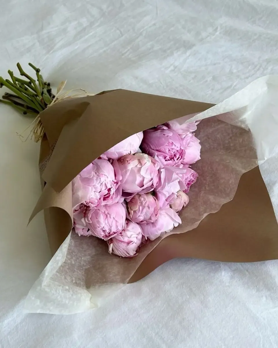 Bó mẫu đơn hồng tím gói giấy kraft và giấy trắng mỏng, đặt trên nền vải trắng