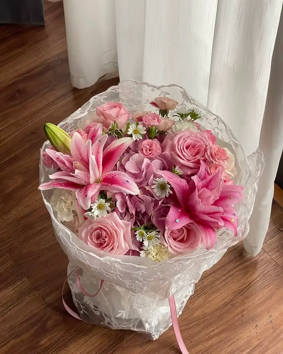 Bó hoa hồng và hoa ly hồng gói trong lớp giấy bóng kính trong suốt đặt trên sàn gỗ cạnh rèm trắng