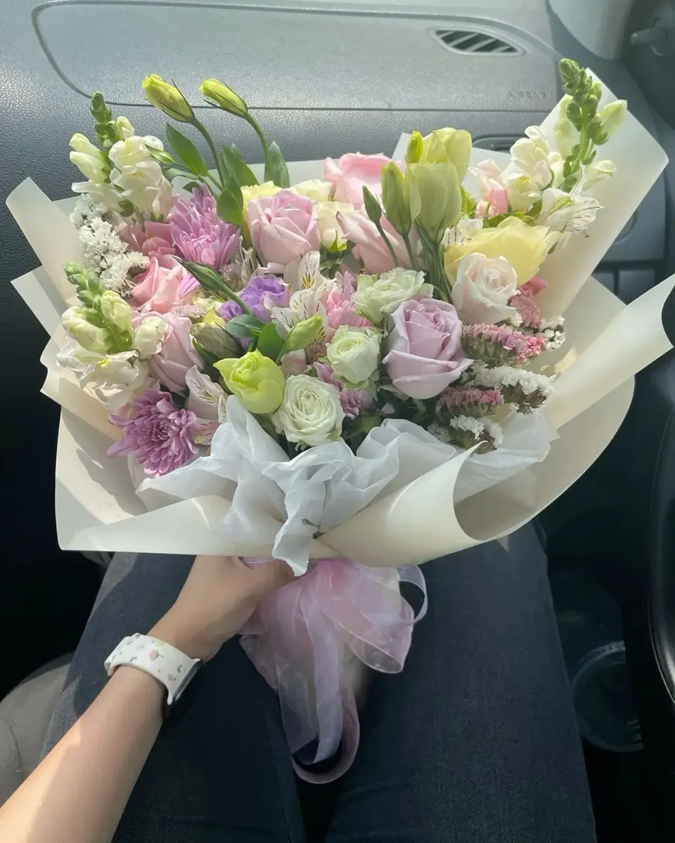 Bó hoa pastel tím hồng và lá xanh gói giấy trắng đặt trên ghế xe