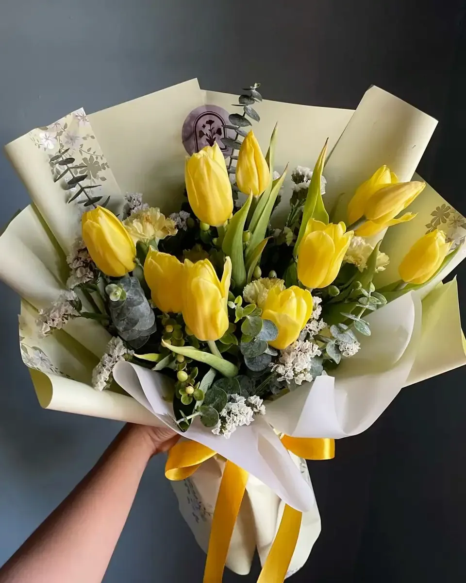 Bó tulip vàng gói giấy kem, buộc ruy băng vàng cầm trên tay trước nền tường tối