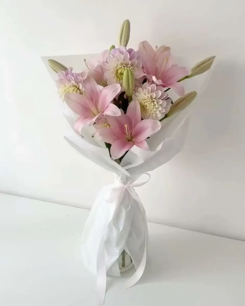 Bó hoa ly hồng nhạt và hoa tím nhạt gói giấy trắng, đặt trên mặt bàn trắng