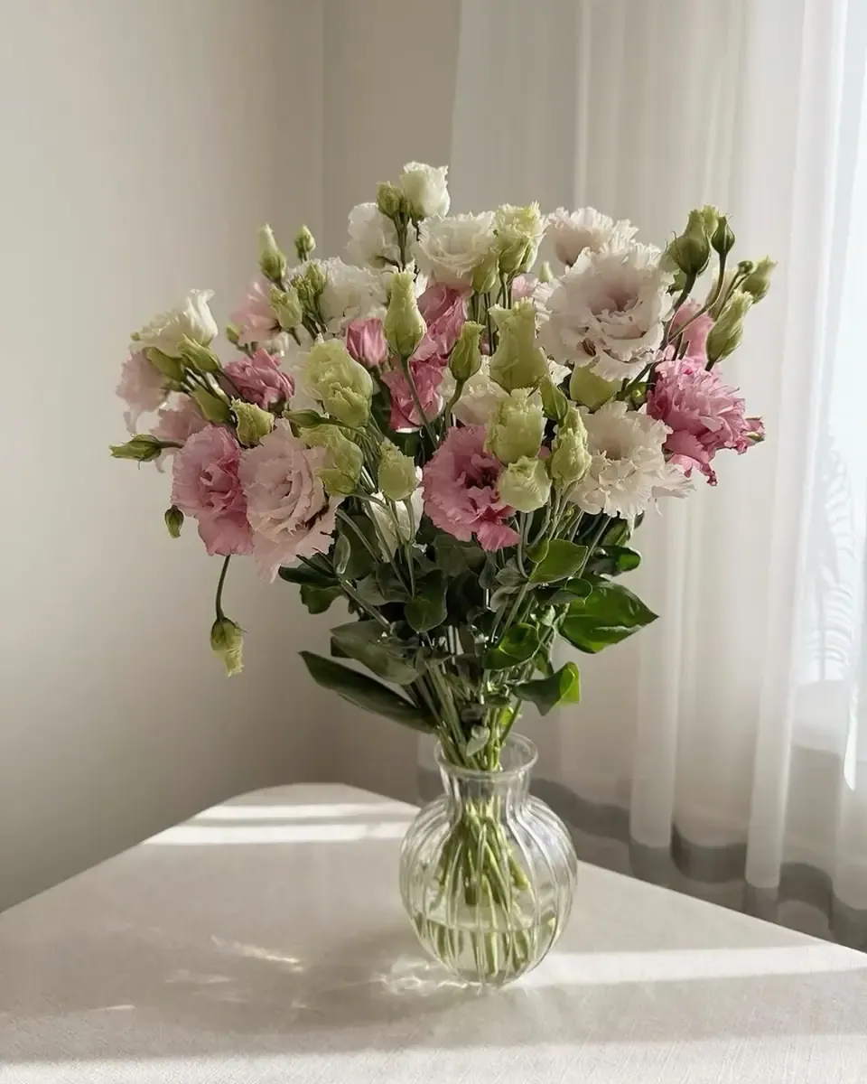 Bình thủy tinh nhỏ cắm cát tường hồng, trắng và nụ xanh trên bàn trắng cạnh rèm voan