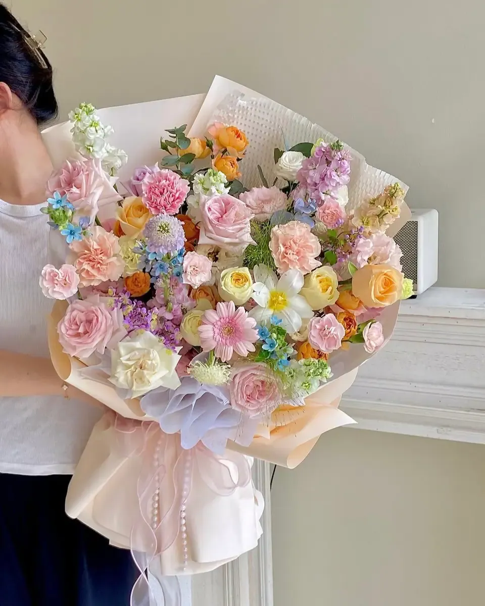 Bó hoa nhiều màu pastel gồm hồng, cam, tím và xanh, được ôm trước một bức tường sáng