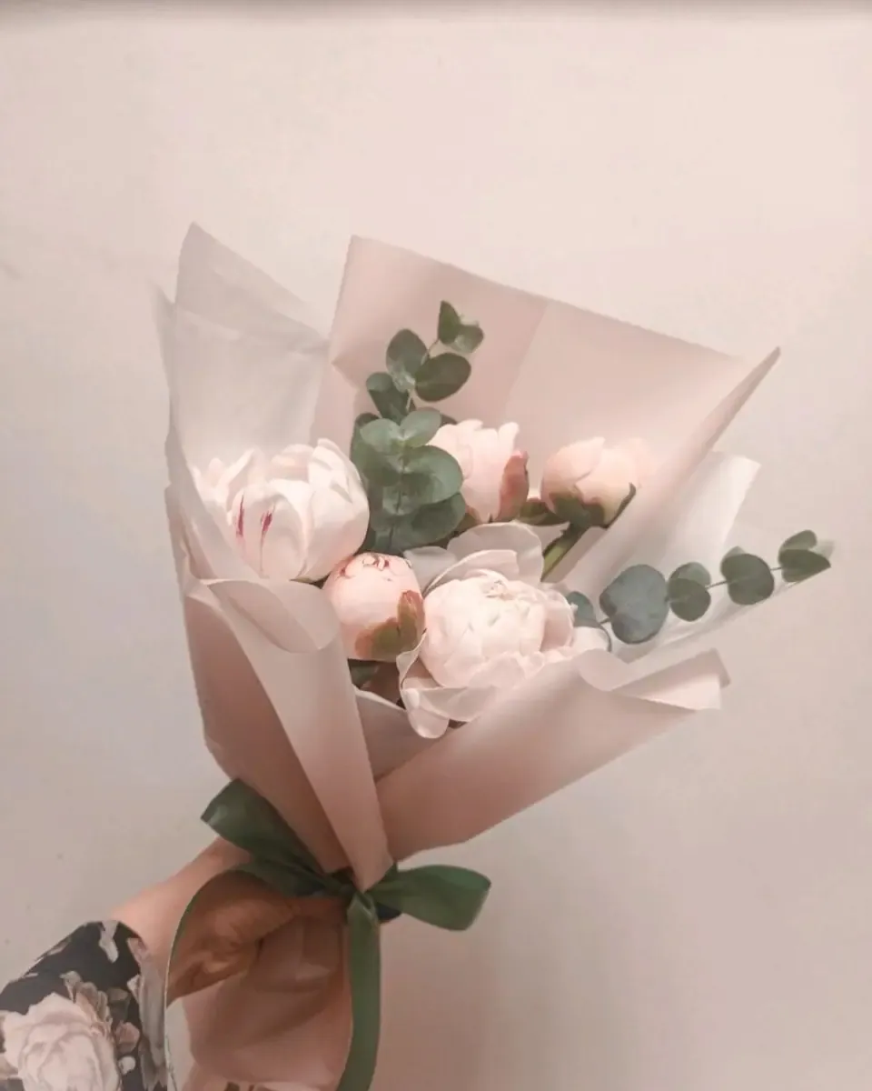 Bó mẫu đơn hồng và trắng nhỏ gói giấy trắng ngà, buộc ruy băng xanh rêu, được cầm trên tay