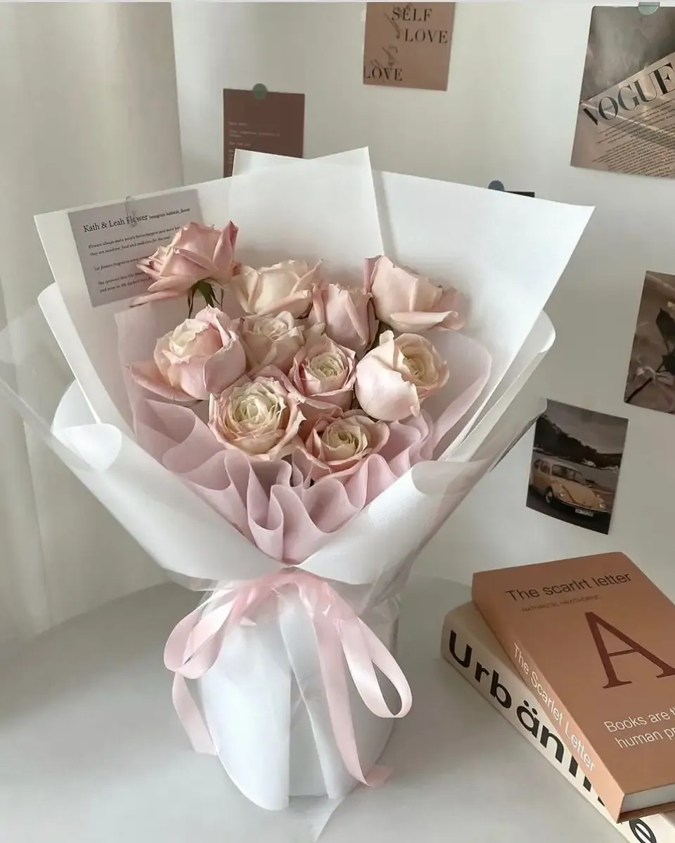 Bó hoa hồng phấn gói giấy trắng và hồng, buộc ruy băng hồng, đặt trên bàn cạnh sách
