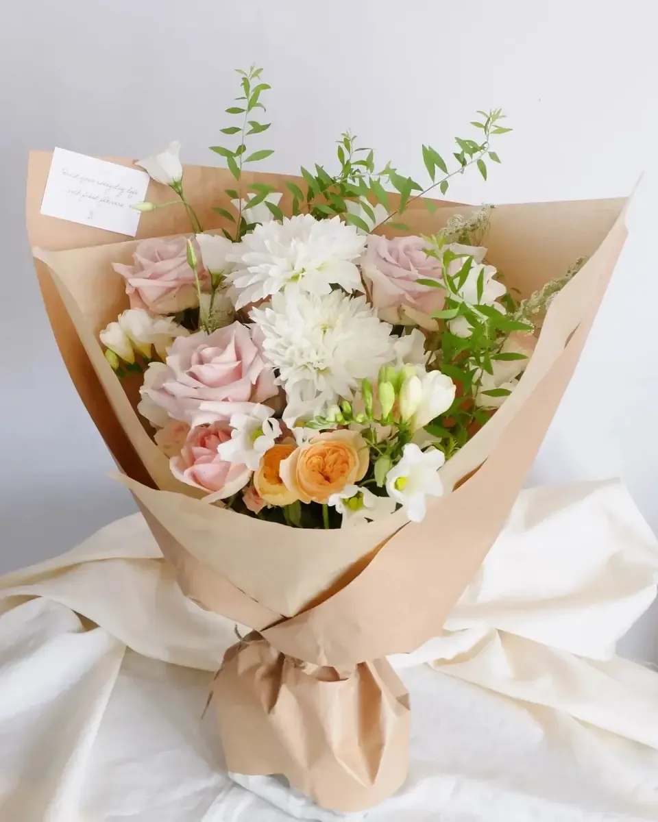Bó hồng phấn nhỏ cùng cúc trắng và hồng cam gói giấy kraft đặt trên vải trắng