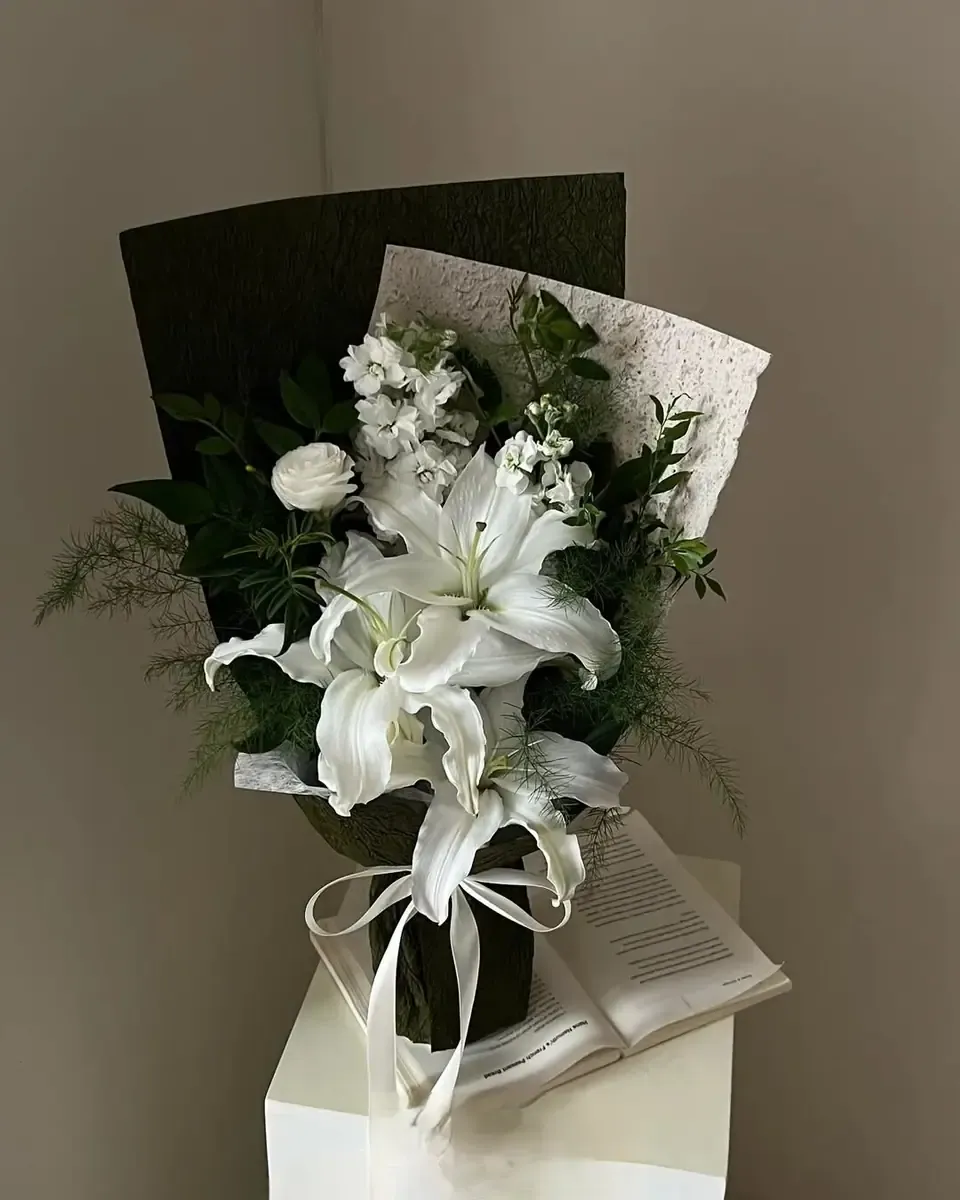 Một nhành ly trắng cùng lá xanh gói giấy xám và đen, đặt trên bục trắng cạnh cuốn sách