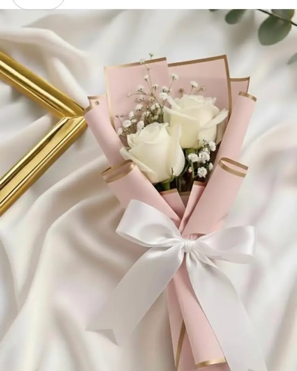 Bó hoa nhỏ hồng trắng gói giấy hồng viền vàng, buộc nơ trắng, đặt trên khăn lụa kem
