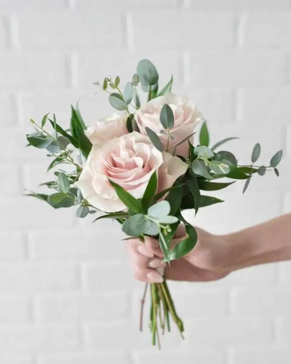 Bó hoa hồng phấn, lá bạch đàn đưa trao bằng tay trước tường gạch trắng