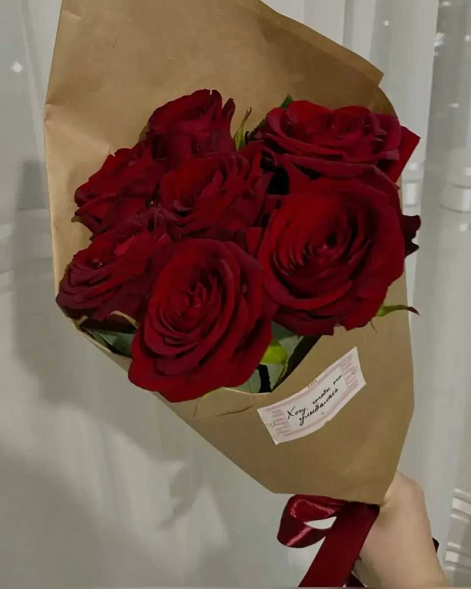 Hoa hồng đỏ sẫm gói giấy kraft có nhãn nhỏ, người cầm giữ bó hoa cạnh dải ruy băng đỏ