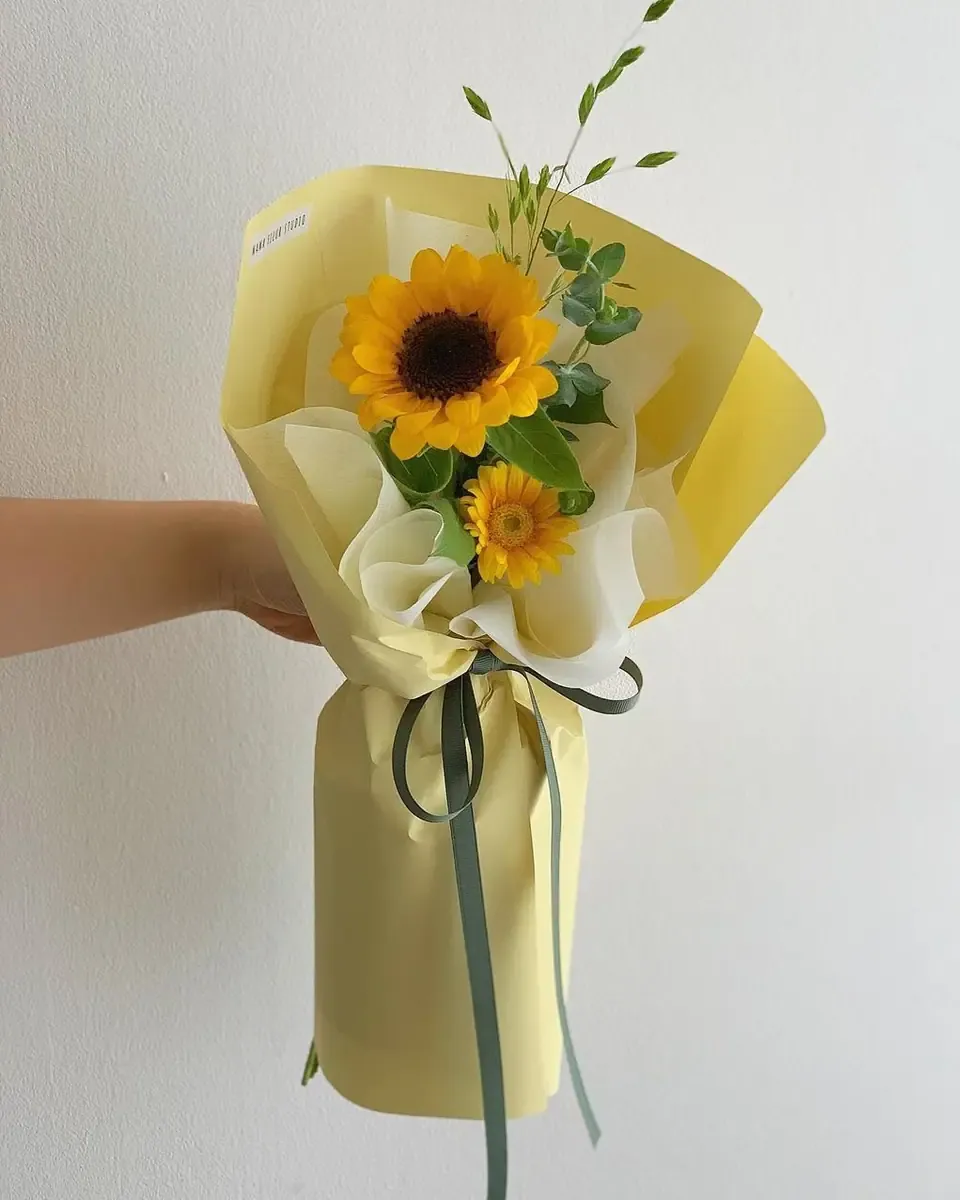 Bó hướng dương nhỏ gói giấy vàng nhạt, buộc ruy băng xanh rêu, được cầm trên tay