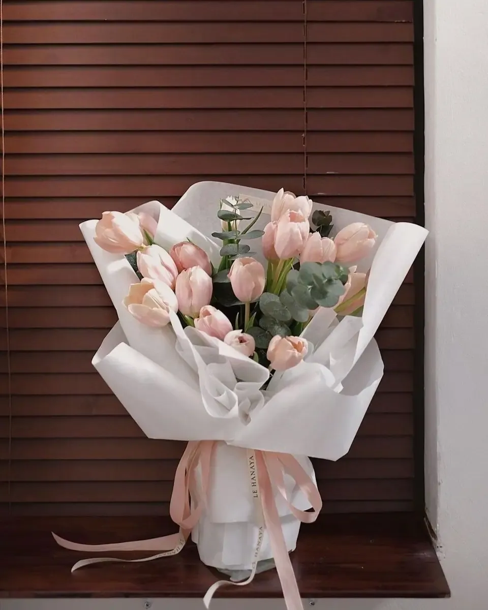 Bó tulip hồng gói giấy trắng và lá bạch đàn, buộc ruy băng hồng đặt cạnh cửa chớp gỗ