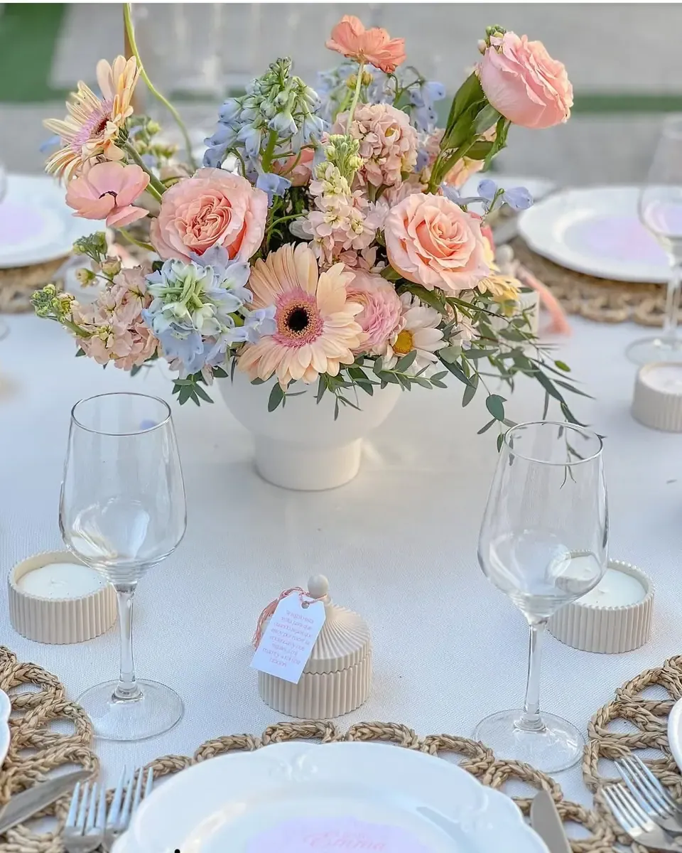 Bình hoa hồng cam, gerbera và hoa nhỏ đặt trên bàn tiệc với ly và đĩa mây tre