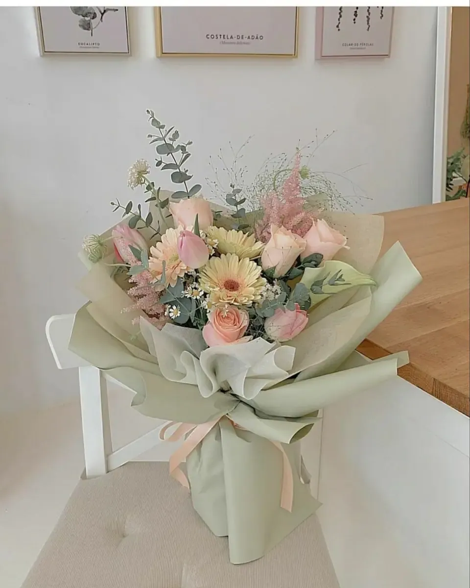 Bó hoa pastel cam hồng kem gói giấy xanh sage, đặt trên ghế gỗ gần tường sáng