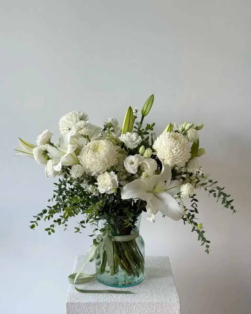 Bình thủy tinh buộc dây cắm cúc trắng, hoa ly và lá xanh, đặt trên bục trắng trước tường sáng
