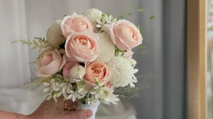 Bàn tay cầm bó hoa hồng đào và hoa trắng nhỏ trước tấm gương, nền rèm xám dịu
