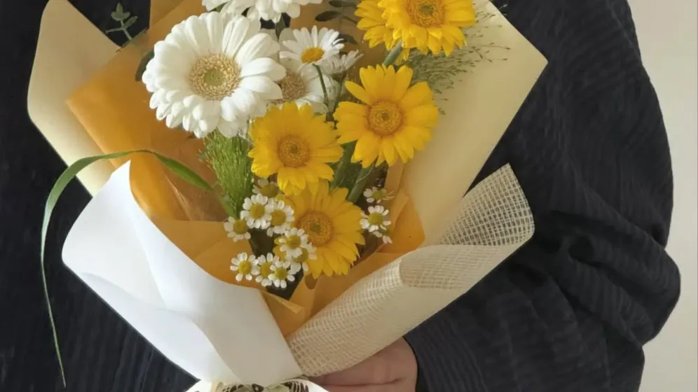 Bó gerbera vàng và cúc trắng gói giấy kem trong tay người mặc áo tối