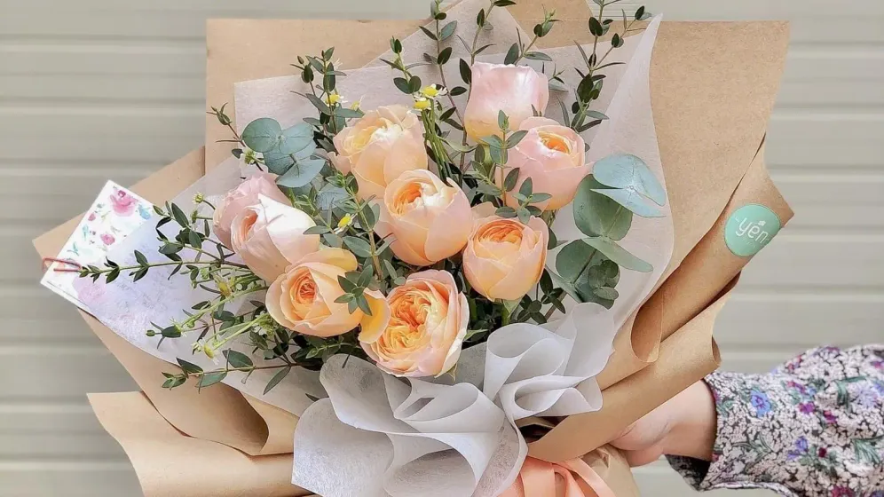 Bó hồng cam đào xen lá bạch đàn gói giấy kraft và giấy kem, buộc ruy băng cam, một tay cầm