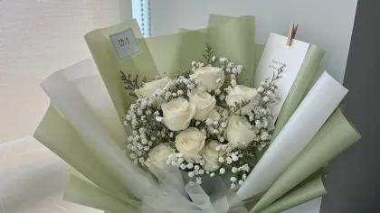 Bó hoa trắng gồm hồng, cúc và lá bạc hà gói giấy xanh sage, đặt cạnh cửa sổ