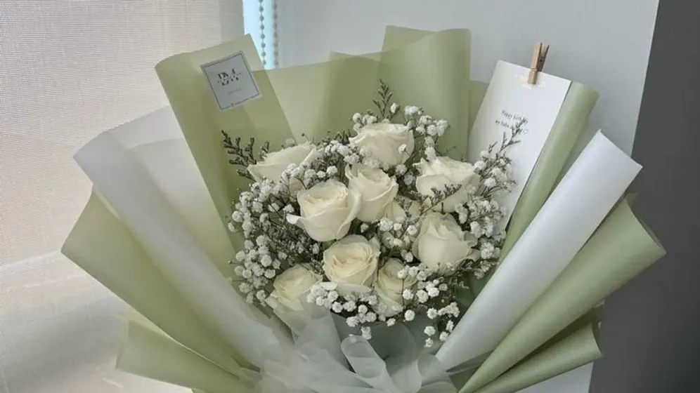 Bó hoa trắng gồm hồng, cúc và lá bạc hà gói giấy xanh sage, đặt cạnh cửa sổ
