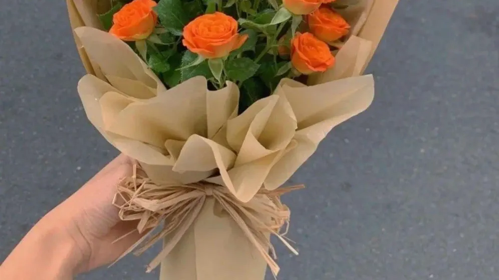 Bó hồng cam nhỏ gói giấy be, buộc dây rơm, được cầm trên tay