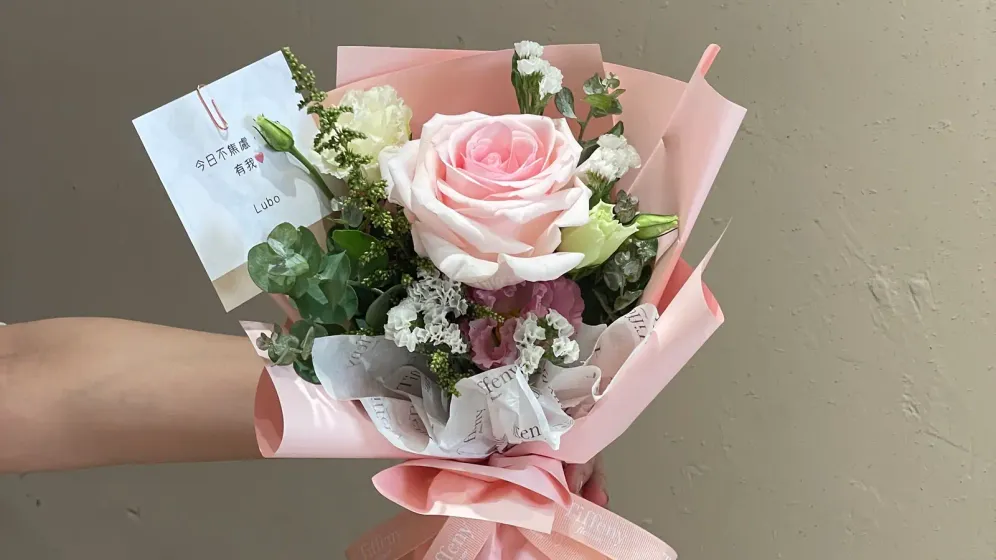 Bó hoa nhỏ tông hồng gói giấy hồng nhạt, được cầm trên tay trước bức tường màu be