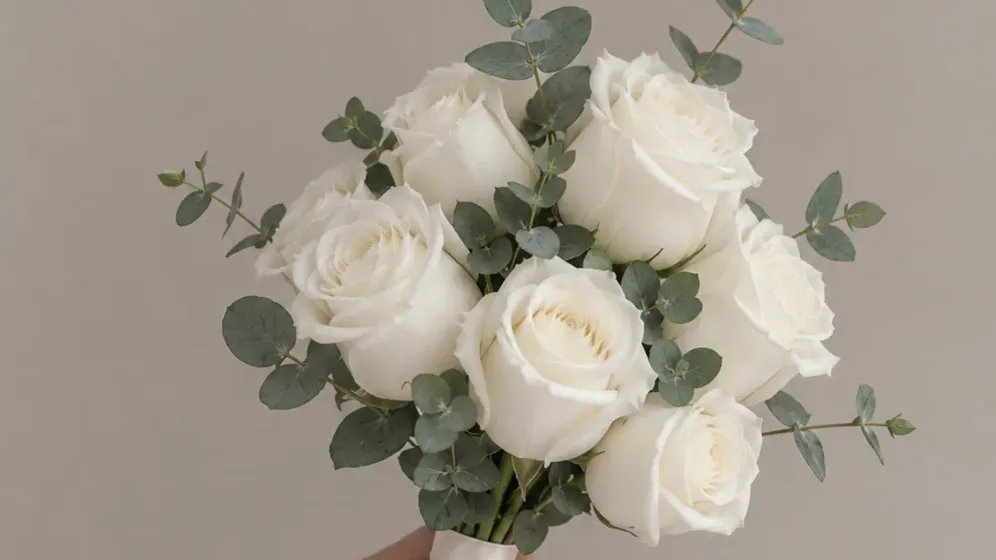 Bàn tay cầm bó hoa hồng trắng kèm lá bạch đàn xanh bạc trên nền xám sáng