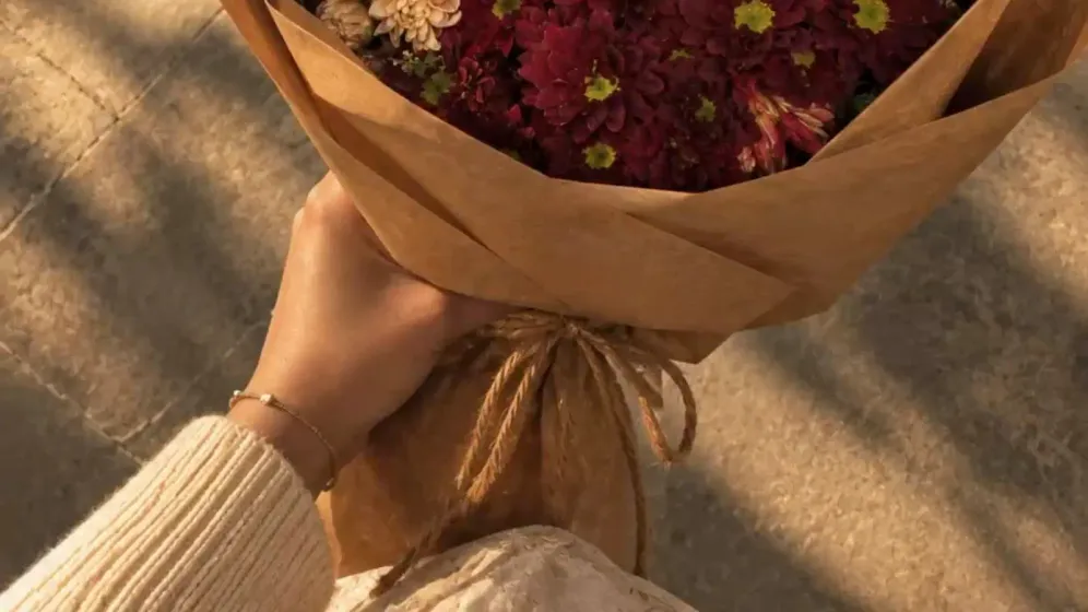 Bó cúc hồng đào và đỏ đô gói giấy kraft nâu, cầm trên đường nắng chiều