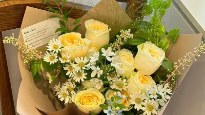 Bó hồng kem và cúc họa mi gói giấy kraft, đặt trên ghế gỗ cạnh cửa sổ