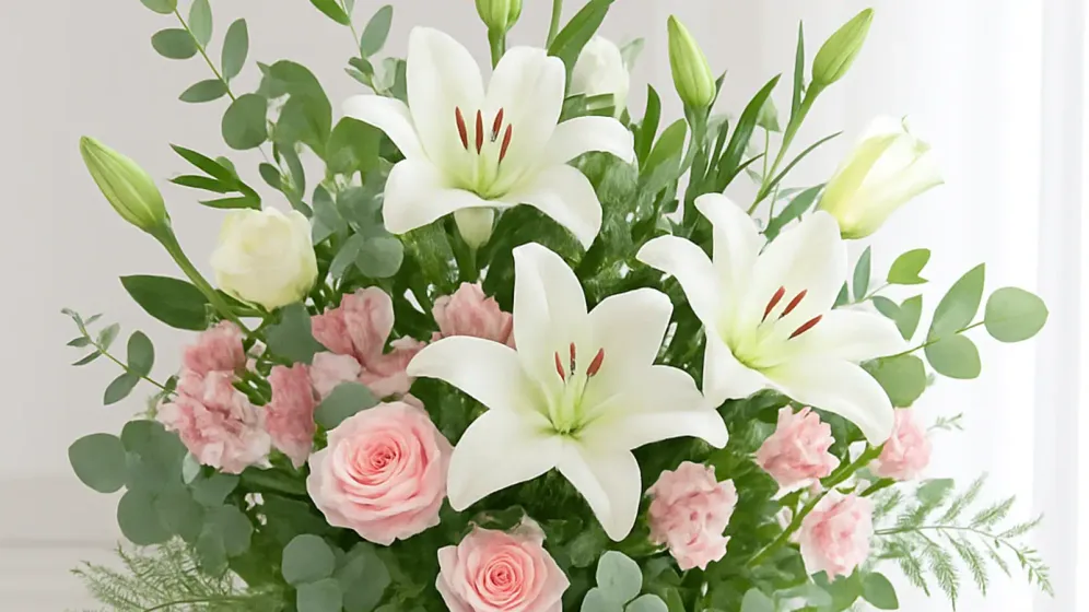 Bình thủy tinh cắm ly trắng, hồng phấn và lá bạch đàn đặt trên bàn trắng cạnh rèm sáng
