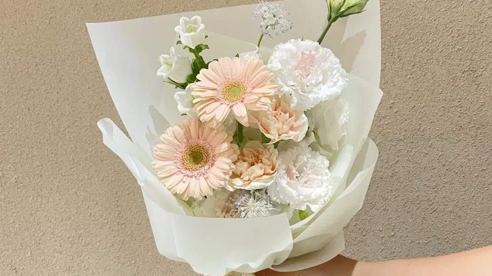 Bó hoa nhỏ tông kem, hồng đào gói giấy trắng, buộc ruy băng be, một tay cầm