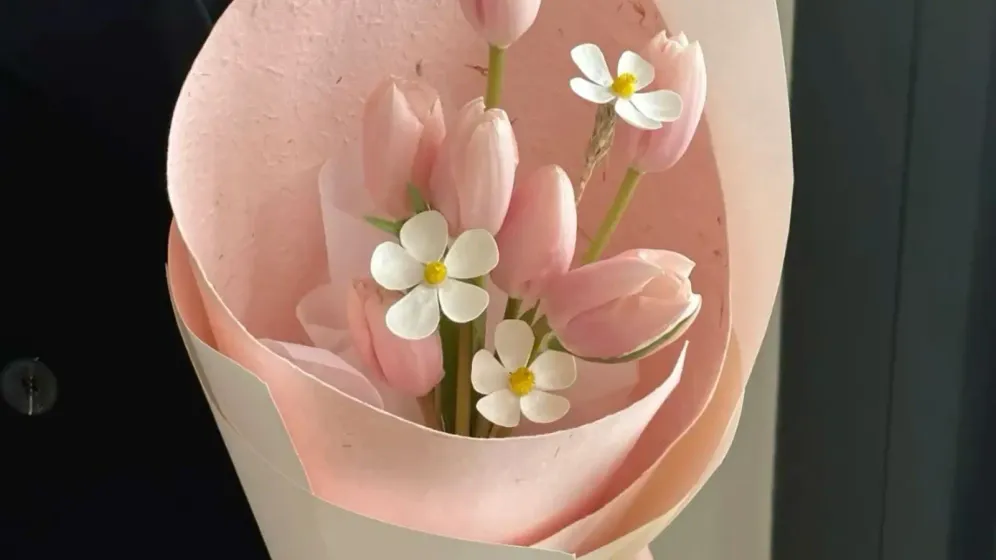 Bó tulip hồng nhạt gói giấy hồng phấn, thêm vài bông hoa trắng nhỏ, một tay cầm bó hoa