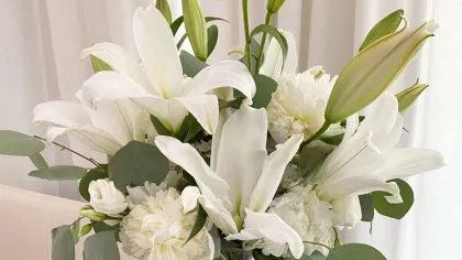 Bình thủy tinh thấp cắm hoa ly trắng, cẩm chướng trắng và lá bạc hà đặt cạnh rèm cửa sáng